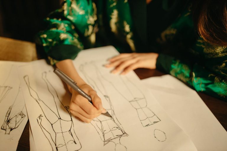 O que a trajetória de Vera Wang revela sobre construir uma marca de moda nos EUA Designer esboça uma peça de roupa em um caderno dentro de um ateliê de moda.